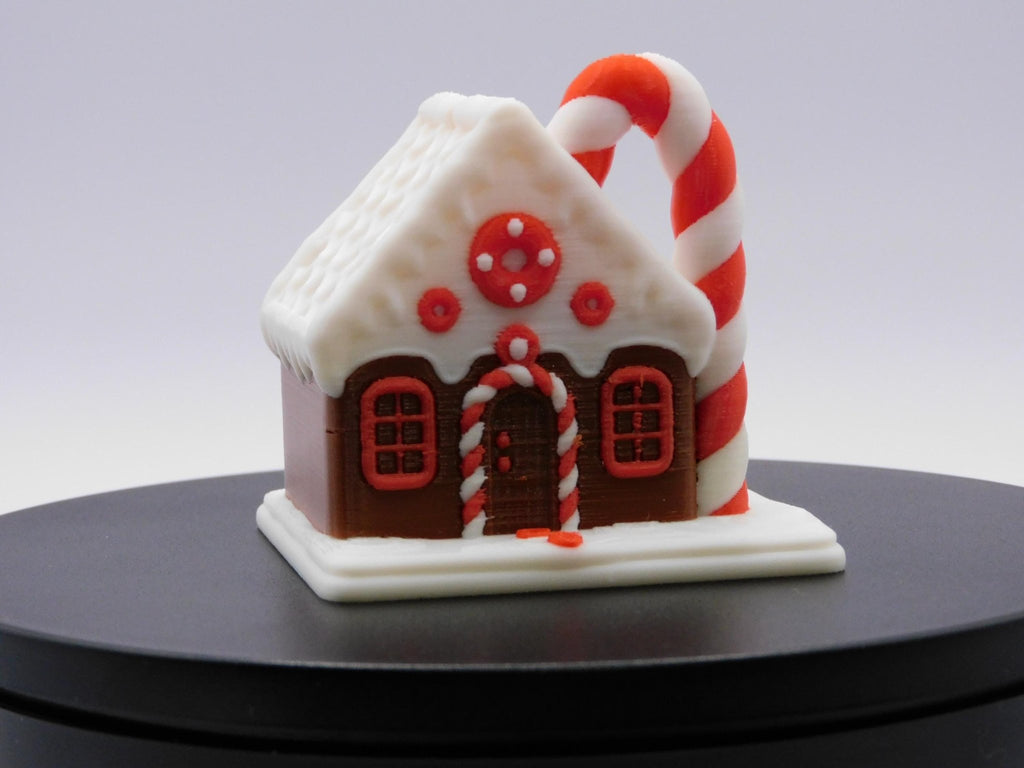 3D printed gingerbread house ornament with a white icing-style roof and red candy decorations.- JM Designs